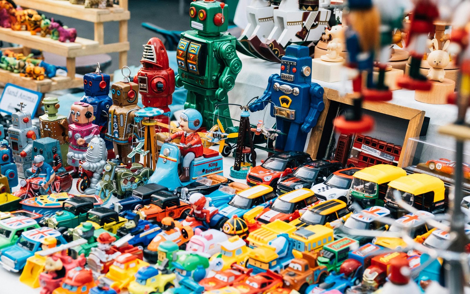 A close-up, high-angle view of a large, colorful collection of vintage and retro toys displayed on a table. The display is dominated by metal and plastic toy cars, trucks, and several brightly colored toy robots (blue, green, and red) standing upright in the background.